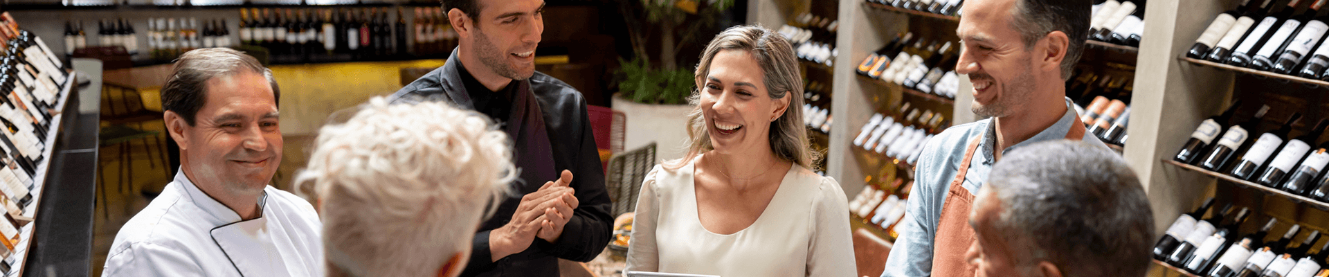 Multiple co workers talking to each other in a meeting in a restaurant surrounded by wine bottles.