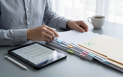 Man's hands on desk with tablet and papers, including draft and final versions. Coffee cup nearby. Focus on business documentation, review, and workflow management in an office setting.